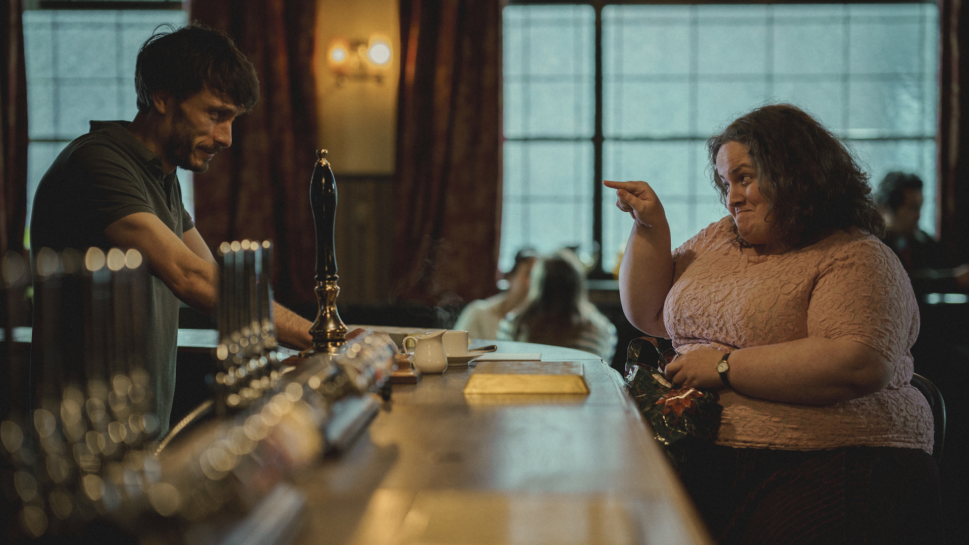 A male bartender stands across the bar from a female patron pointing at him with a smile; still from 'Baby Reindeer'