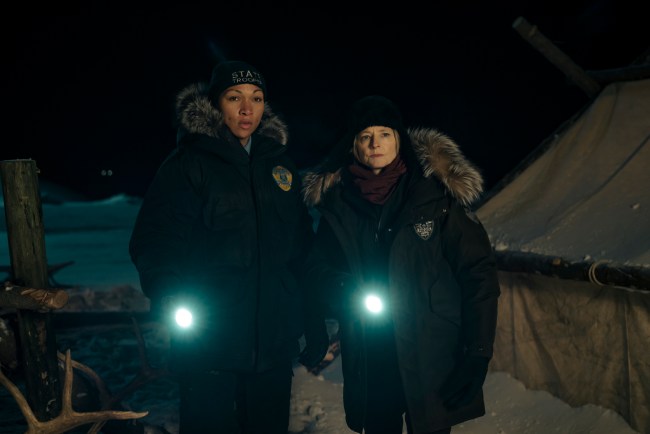 Two women in thick winter coats and hats holding flashlights while out in the snow at night; still from "True Detective: Night Country"