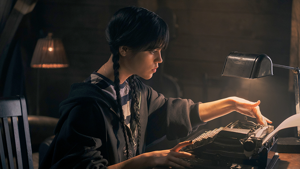 Wednesday (Jenna Ortega) sitting at her typewriter in her dorm room, gazing a typed sheet of paper in "Wednesday"