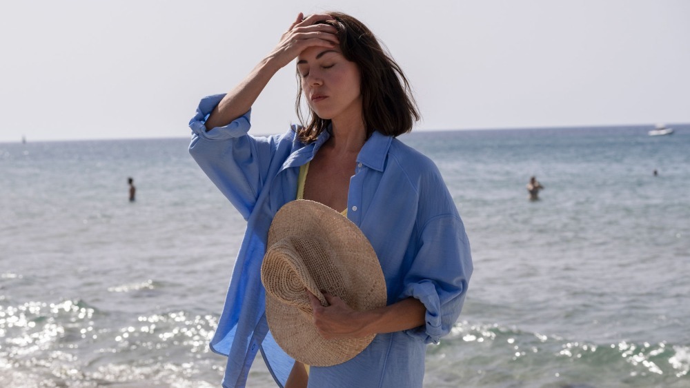 Aubrey Plaza in "The White Lotus" as Harper, standing in beach clothes in front of the ocean, hand on head and eyes closed like she has a headache.