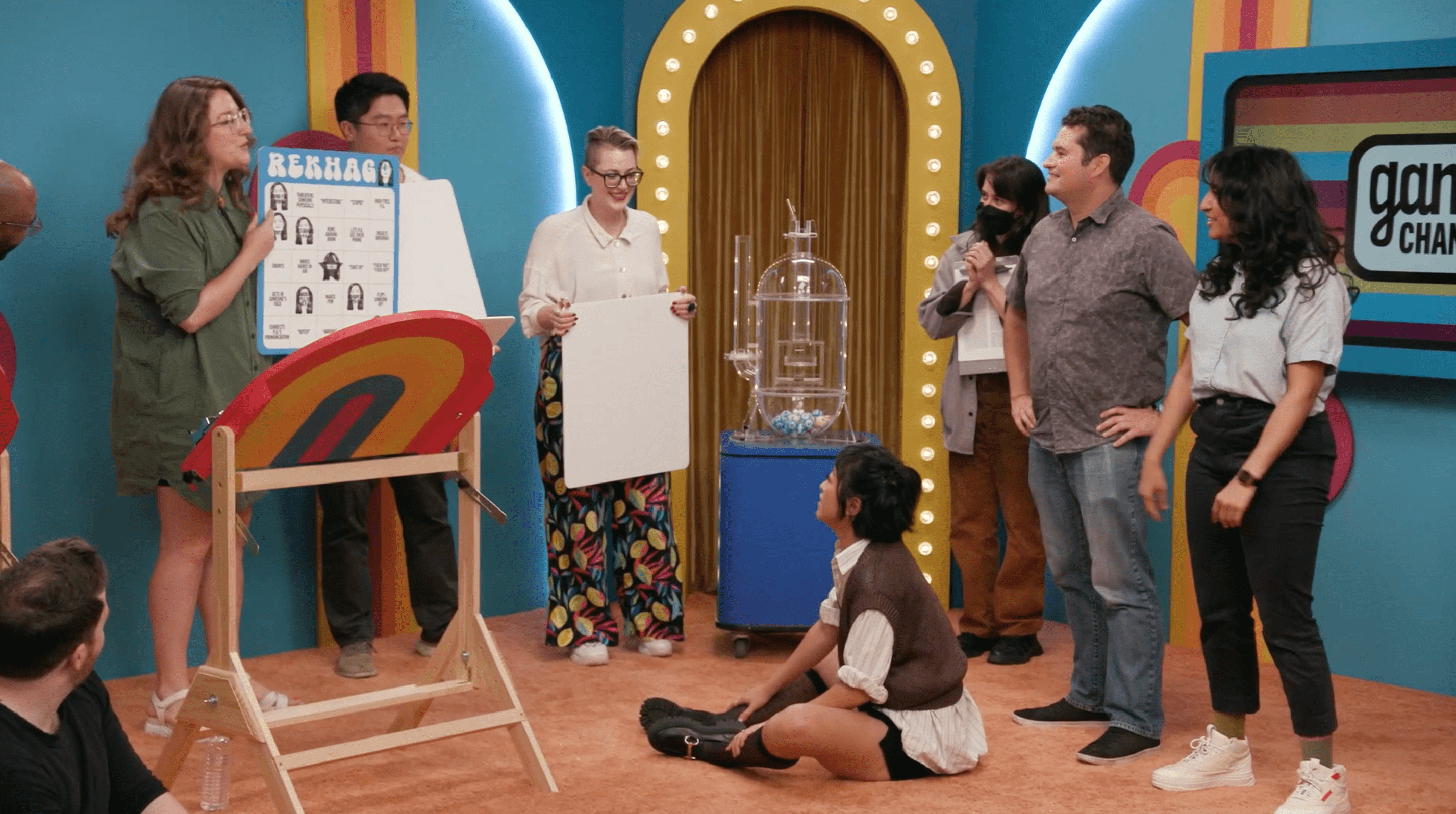 The cast of the 'Game Changer' episode 'Bingo' gather on set around Jessica Ross holding a large blue and white bingo board that says 'Rekhago.' From left to right: Brennan Lee Mulligan, Raphael Chestang, Jessica Ross, Tao Yang, Carolyn Page, Lily Du, Kaylin Mahoney, Mike Trapp, Rekha Shankar